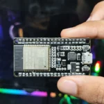 Hình ảnh một người đang cầm module vi điều khiển ESP32-WROOM-32 trước màn hình máy tính